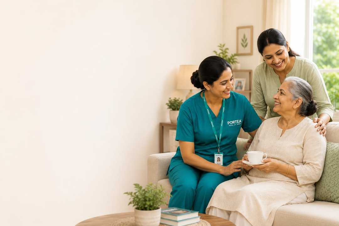 A Portea caregiver in a teal uniform sitting with an elderly woman holding a cup of tea on a sofa, while a younger woman stands behind them smiling